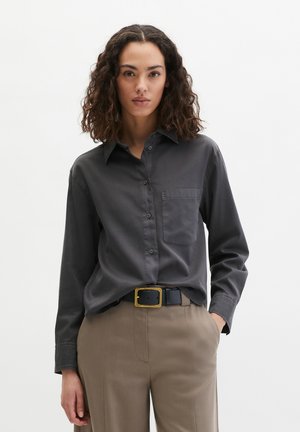 Frau mit lockigem Haar, die ein dunkelgraues Langarmshirt trägt, das in beige Hosen mit einem schwarzen Gürtel und goldener Schnalle gesteckt ist, steht vor weißem Hintergrund.