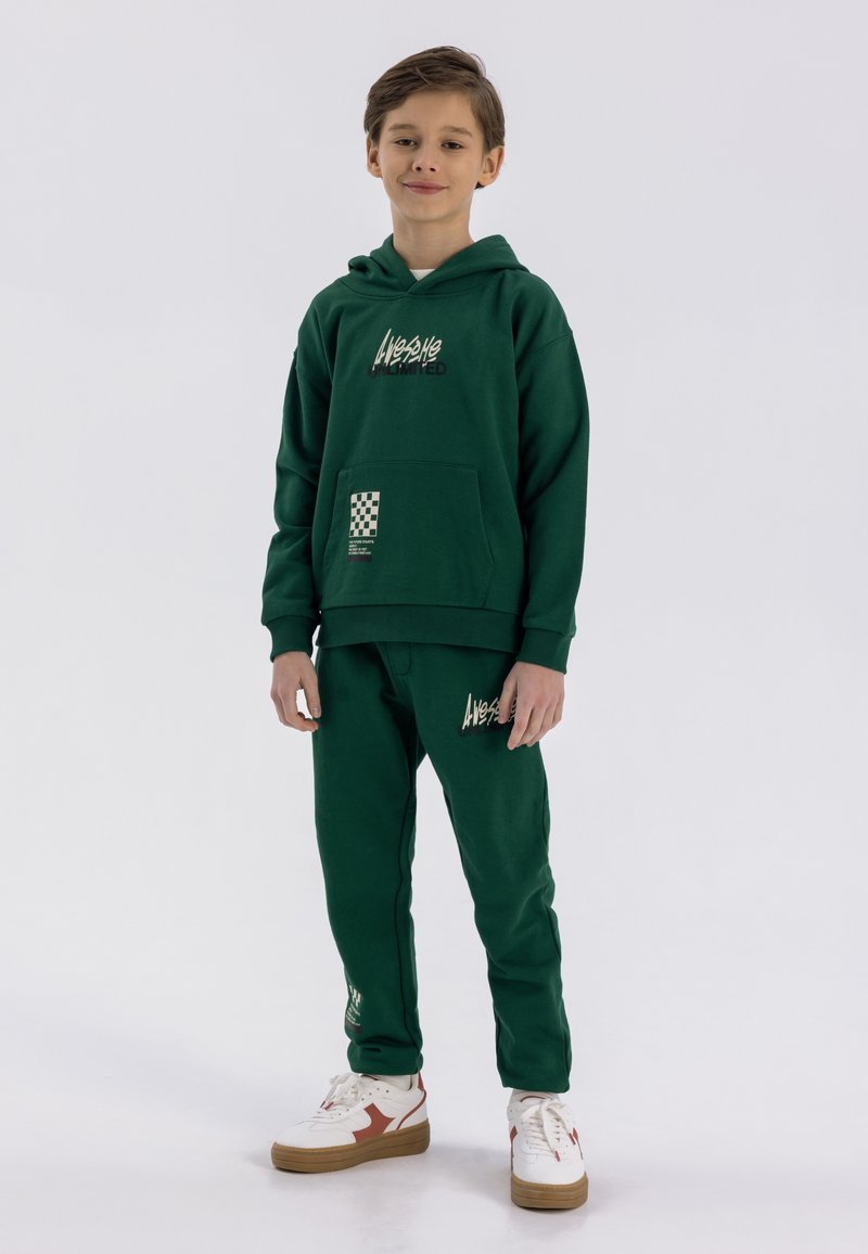 Zaļš hoodie ar priekšējo kabatu un baltām logo, pārī ar atbilstošām sporta biksēm. Akcenti ietver rūtainu motīvu kabatā. Baltas kedas.