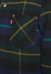 Camisa de franela con un estampado de cuadros en verde, negro y púrpura, con rayas amarillas, un bolsillo en el pecho y una etiqueta de marca roja.