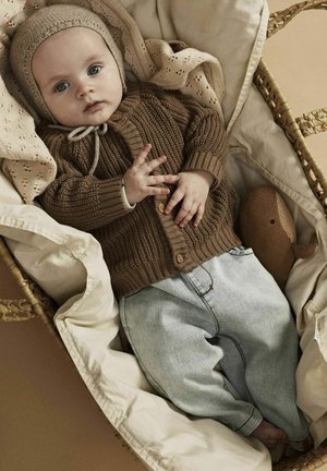 Brauner Strickpullover mit Knöpfen, hellblaue Jeanshose und beiger Strickmütze, die auf einer cremefarbenen Decke in einem geflochtenen Korb ruhen.