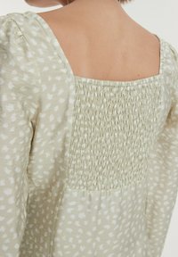 Hellgrünes, gemustertes Kleid mit quadratischem Rückendesign. Verfügt über Rüschendetails entlang des Rückens und einen weichen, fließenden Stoff mit subtilen Texturen.