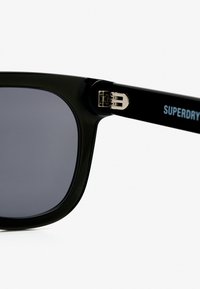 Czarne okulary przeciwsłoneczne o prostokątnych soczewkach, z metalowymi srebrnymi akcentami i błyszczącym wykończeniem. Na ramieniu widnieje nazwa marki "SUPERDRY" w kolorze niebieskim.