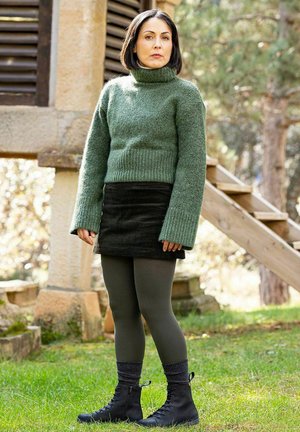 Mujer de pie al aire libre cerca de un edificio de piedra y escaleras de madera, vistiendo un suéter verde de cuello alto, falda oscura, leggings y botas negras.