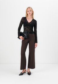 Mujer vestida con un top negro de encaje de manga larga, pantalones anchos oscuros, tacones negros puntiagudos, sosteniendo un bolso de cuero negro.