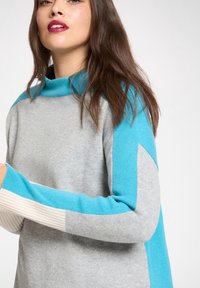 Grauer Pullover mit türkisfarbenen Akzentärmeln und hohem Kragen. Verfügt über gerippte Texturen und ein Farbblockdesign. Weiches Strickmaterial.