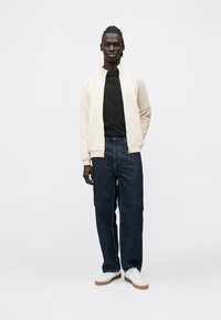 Lys beige bomberjakke med glidelås over en svart T-skjorte, kombinert med mørkeblå vide jeans og hvite joggesko med brune detaljer.