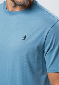 Homme portant un t-shirt bleu clair à col rond avec une petite figure sombre brodée sur le côté gauche de la poitrine, manches courtes, et une barbe visible.