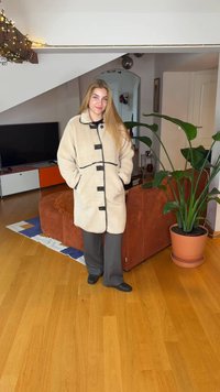 Krämfärgad fleecemantel med svarta detaljer, knappstängning och sidofickor. Bärs över grå byxor, kombinerat med svarta skor. Inomhusmiljö.