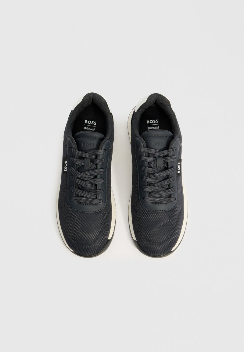 Une paire de baskets BOSS noires avec une texture en maille, un design à lacets et des semelles blanches rembourrées sur un fond gris clair.