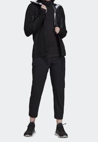 Veste zippée noire en tissu texturé, associée à un pantalon noir fuselé et des chaussures de sport noires avec des accents gris.