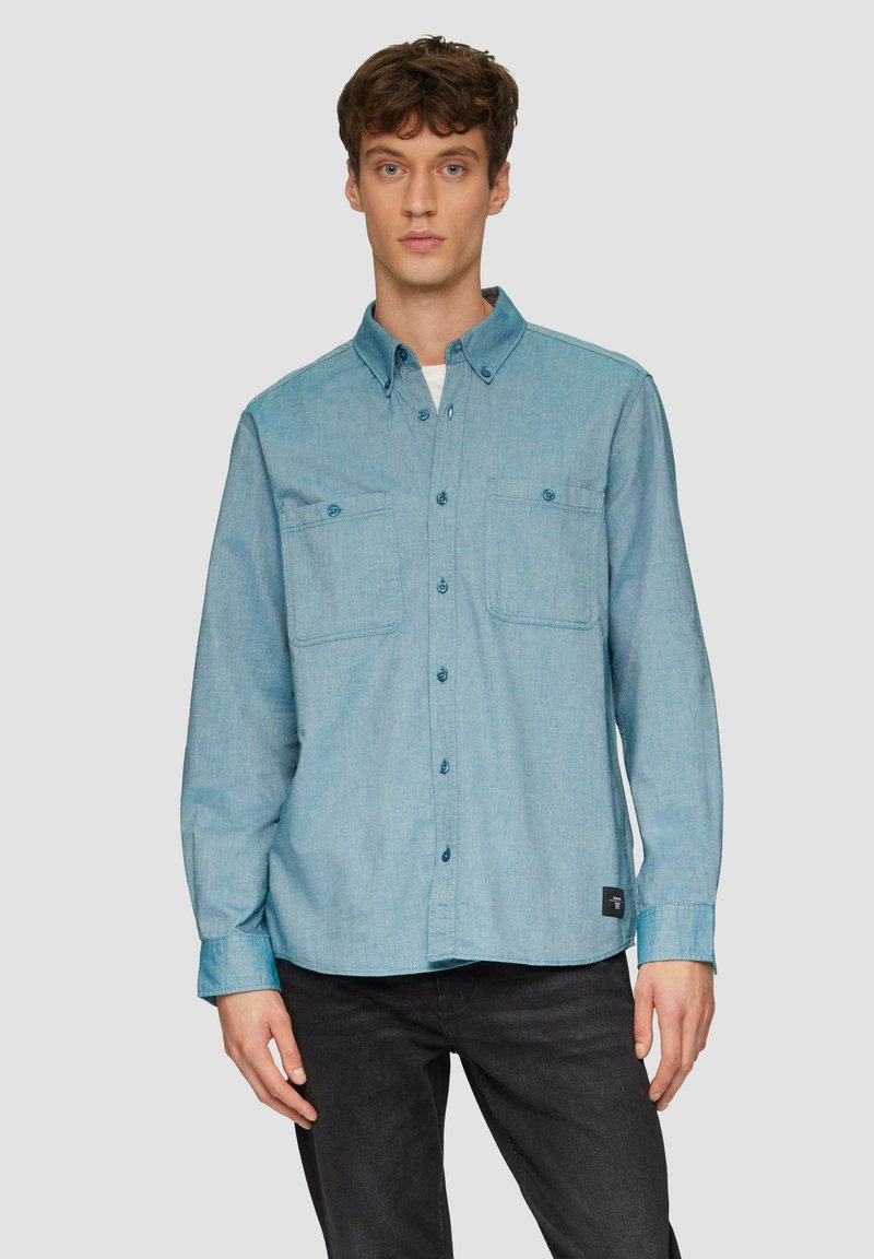 Lichtblauw overhemd met knopen, gemaakt van denim, met twee borstzakken, lange mouwen en een ronde zoom. Gestyled met een donkere denim achtergrond.