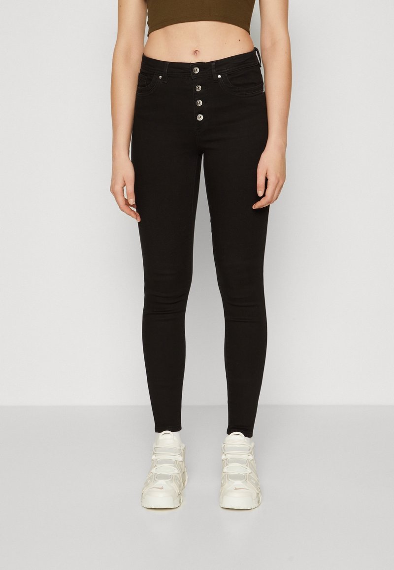 Jeans skiny negros con cintura alta, cierre de cinco botones y bolsillos tradicionales, combinados con zapatillas deportivas blancas. Textura de denim suave.