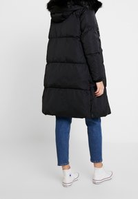 Abrigo acolchado negro con capucha y ribete de piel sintética, con diseño acolchado, corte hasta la rodilla y bolsillos laterales con cremallera. Se lleva sobre vaqueros azules.