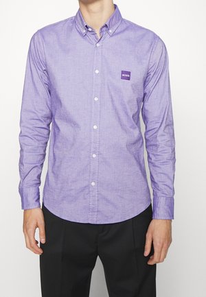 Mężczyzna noszący lawendową koszulę z długim rękawem zapinaną na guziki z naszywką z logo „BOSS” na lewej piersi, w połączeniu z czarnymi spodniami.