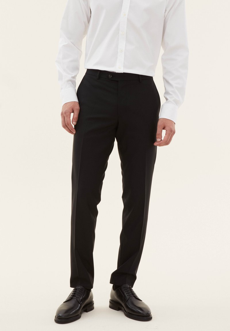 Pantaloni neri su misura realizzati in tessuto morbido, con una vestibilità slim, passanti per cintura e chiusura con un solo bottone, abbinati a scarpe eleganti nere.
