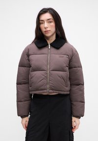 Brązowa krótka kurtka puchowa z czarnym kołnierzem z sztucznego futra, pikowany wzór, srebrny zamek błyskawiczny i haftowane logo po lewej stronie klatki piersiowej.