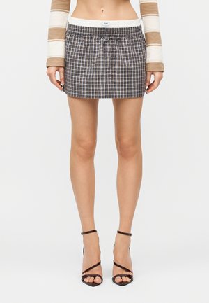 Kobieta ubrana w brązowo-biały pasiasty top z długim rękawem, niebiesko-szarą kratowaną mini spódniczkę oraz czarne sandały na obcasie z paskami.