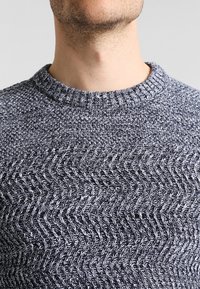 Szary sweter dziany o fakturze, zdobiony wzorem w jodełkę, z okrągłym dekoltem i ściągaczami przy rękawach. Materiał ma miękkie, tkane wykończenie.