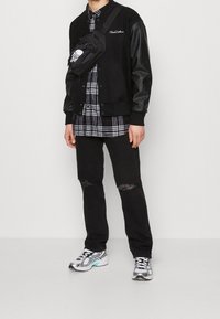 Czarna kurtka varsity z skórzanymi rękawami, koszula w kratę pod spodem, przetarte czarne dżinsy i srebrne buty sportowe z akcentami w kolorze teal. Regulowana czarna torba.