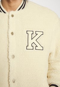 Cream-colored shearling jacket with a letter 'K' patch, snap buttons, and ribbed black and white collar and cuffs. Soft, textured fabric.
