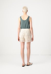 Grøn ærmeløs top, beige blonde shorts med bølgemønster forneden og lyse sandaler. Outfittet har en afslappet pasform og detaljeret blondearbejde.
