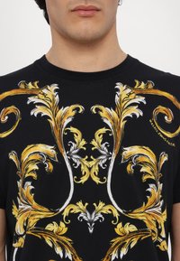 T-shirt preta com intrincados padrões florais barrocos em dourado e branco. Apresenta um decote arredondado e mangas curtas.