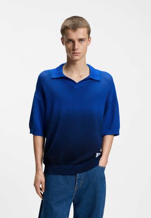 Junger Mann trägt ein blaues, verlaufenes Strick-Poloshirt mit kurzen Ärmeln und blaue Jeans, steht vor einem schlichten weißen Hintergrund.