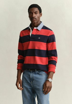 Mann trägt ein rot-navy gestreiftes Rugbyshirt mit hellblauem Kragen, blaue Jeans und eine silberne Armbanduhr und steht vor einem einfachen Hintergrund.