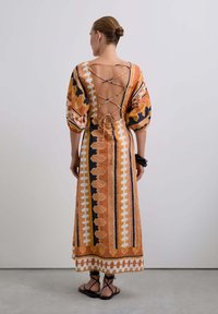 Vue arrière d'une longue robe avec des motifs géométriques orange et noir, des manches bouffantes et un design de dos lacé. Sandales à lanières visibles.