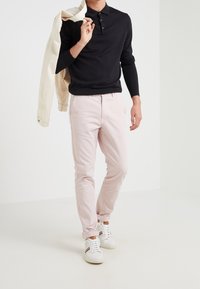 Czarna koszulka polo z długim rękawem, jasnoróżowe chinosy i białe trampki z czerwonymi akcentami. Model niesie beżową kurtkę zarzuconą na jedno ramię.