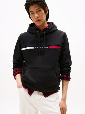 Hoodie en polaire noire avec une poche frontale, ornée d'une bande horizontale blanche et rouge et du logo "TOMMY HILFIGER". Porté par-dessus une chemise à carreaux rouge et noir.