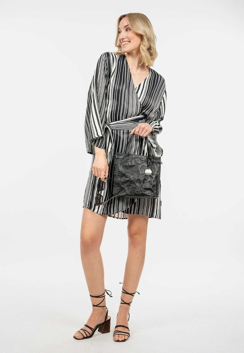 Bolso negro texturizado de forma rectangular, con asas cortas y un acento plateado. La modelo lleva un vestido en forma de envoltura de rayas en blanco y negro y zapatos de tacón con tiras.
