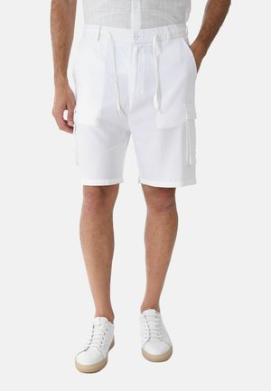 Uomo che indossa pantaloncini cargo bianchi con coulisse e sneakers bianche, in piedi con le mani nelle tasche.