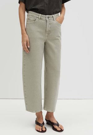 Person trägt hellgraue Jeans mit geradem Bein, ein schwarzes kurzärmeliges Hemd mit Knöpfen und schwarze Wedgesandalen und steht auf einem weißen Boden.