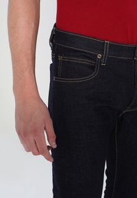 Tmavě modré džíny z denimu s viditelným šitím a pěti kapsami. Látka vypadá odolně a má hladkou texturu. Červený top je částečně viditelný.