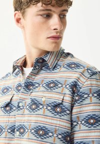 Hemd mit Knöpfen in einem blauen und orangefarbenen geometrischen Muster auf hellem Hintergrund. Hergestellt aus gewebtem Stoff mit zwei Brusttaschen.