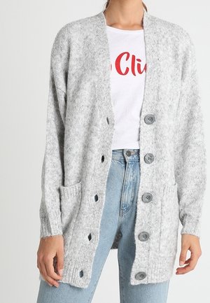 Femme portant un cardigan en laine gris clair avec de gros boutons par-dessus un t-shirt blanc à motif graphique et un jean taille haute bleu clair.