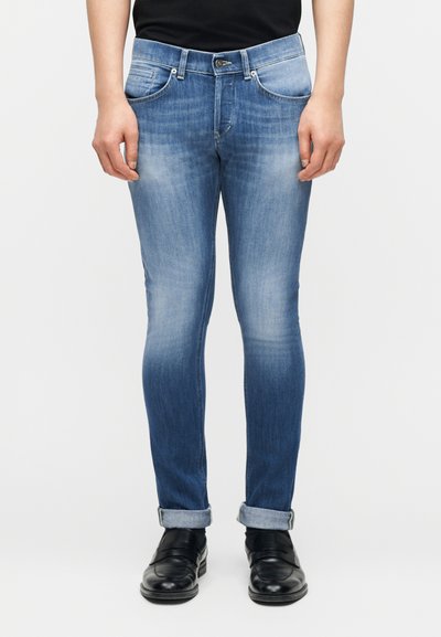 Persoon die opgerolde, slim-fit, vervaagde blauwe jeans draagt met zwarte leren instappers, staand tegen een effen achtergrond.