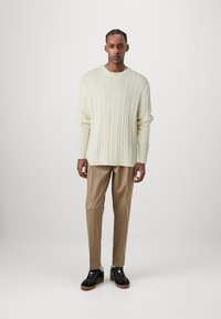 Cream cable-knit sweater with wide cuffs, paired with beige tailored trousers and black sneakers featuring a thick sole. Minimalist design.