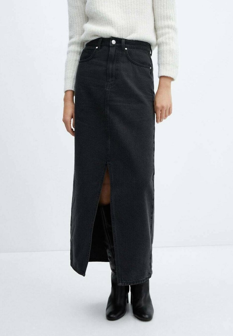 Femme portant une jupe midi en denim noire taille haute avec une fente frontale, un pull blanc en matière pelucheuse et des bottes à talons mi-mollet noires sur un fond blanc.
