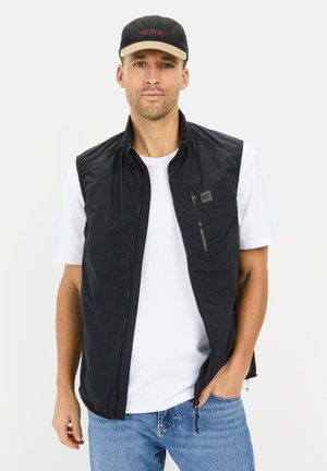 Black zippered vest over a white T-shirt, featuring a small logo and side pockets, worn with light blue jeans and a black-and-tan cap.