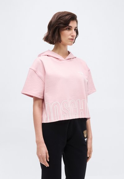 Woman with short brown hair wearing a loose pink hooded t-shirt with large Moschino text and black pants featuring a small teddy bear patch.