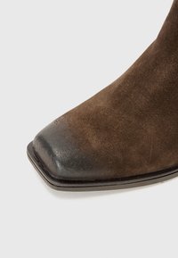 Stivale alla caviglia in suede marrone con punta quadrata, dotato di un elegante cappuccio in pelle nera lucida e suola nera affusolata.