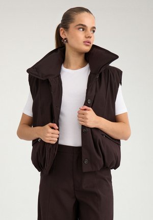 Jeune femme aux cheveux lissés en arrière, portant une veste sans manches matelassée marron foncé par-dessus un haut blanc à manches courtes et un pantalon marron foncé assorti.