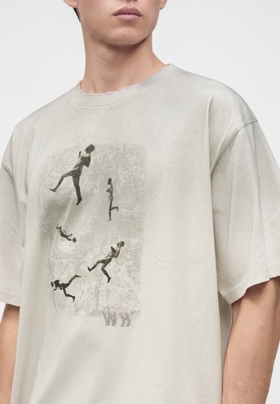 Νέος που φοράει ένα ανοιχτό μπεζ φαρδύ T-shirt με μαύρες αφηρημένες σιλουέτες από δρομείς και άλματα σε κίνηση μπροστά.