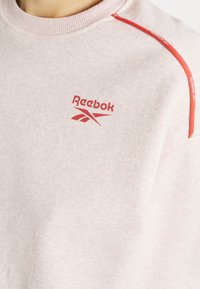 Sweat-shirt rose pâle Reebok avec logo rouge sur la poitrine et passepoil rouge le long de la couture de l'épaule, porté par une personne.