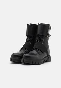 Bottes de combat en cuir noir avec une semelle en caoutchouc épaisse, fermeture à lacets sur le devant et sangles ajustables avec boucles en haut. Texture lisse.