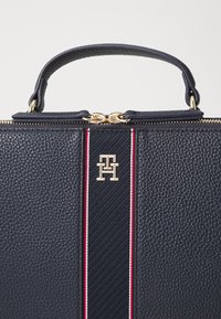 Borsa a tracolla in pelle texturizzata blu navy con logo dorato e una striscia verticale con accenti rossi e bianchi. Due manici superiori e finiture dorate.