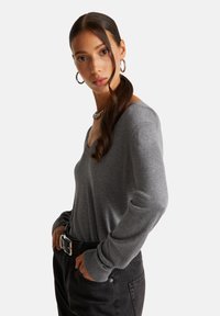 Frau mit langen, dunklen Haaren im Pferdeschwanz, die silberne Creolen, eine dicke Kettenhalskette, ein graues Langarm-V-Ausschnitt-Oberteil und schwarze Jeans mit Gürtel trägt.