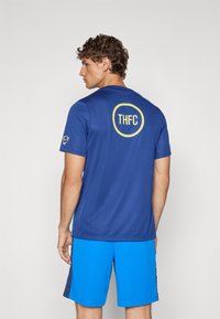 Blå atletisk skjorte med gul "THFC" logo på ryggen, parret med blå shorts, lavet af letvægts, glat materiale.